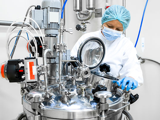 Alt-Text-Vorschlag: &bdquo;Eine Person in Schutzkleidung, Haube, Mundschutz und Handschuhen arbeitet an einem gro&szlig;en Edelstahl-Reaktor in einem Labor oder einer Produktionsanlage.&ldquo;