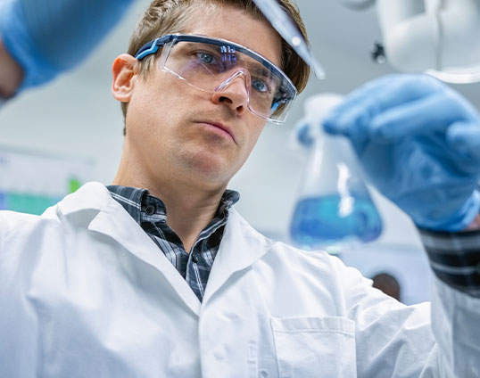 Ein Wissenschaftler im Labor analysiert eine Fl&uuml;ssigkeit in einem Erlenmeyerkolben und tr&auml;gt Schutzbrille, Handschuhe und Laborkittel.