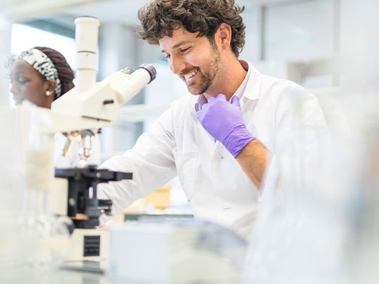 L&auml;chelnder Wissenschaftler arbeitet am Mikroskop, Kollegin im Hintergrund