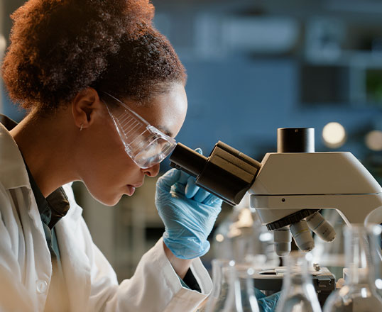 Wissenschaftlerin im Labor schaut durchs Mikroskop, umgeben von Reagenzgl&auml;sern