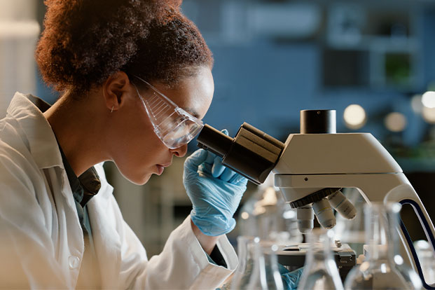 Wissenschaftlerin im Labor schaut durchs Mikroskop, umgeben von Reagenzgl&auml;sern