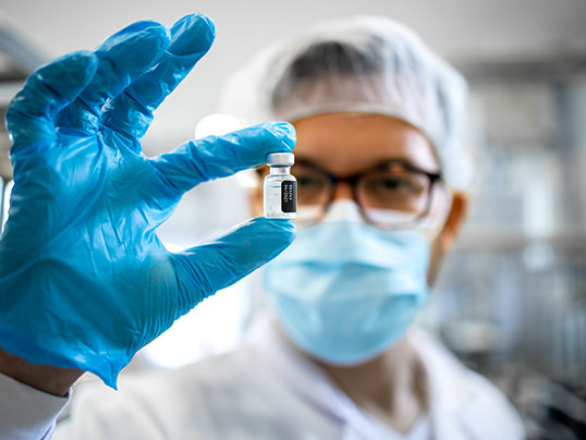 Laborant mit Schutzkleidung h&auml;lt ein Fl&auml;schchen Impfstoff in die Kamera