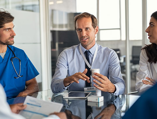 Gesch&auml;ftsmann spricht mit medizinischem Personal, auf dem Tisch Medikamente